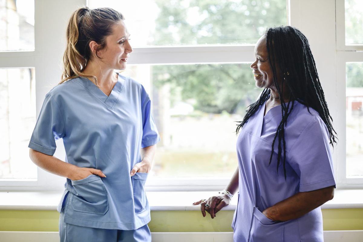 Two care staff in scrubs talking beside a window at the start of a shift