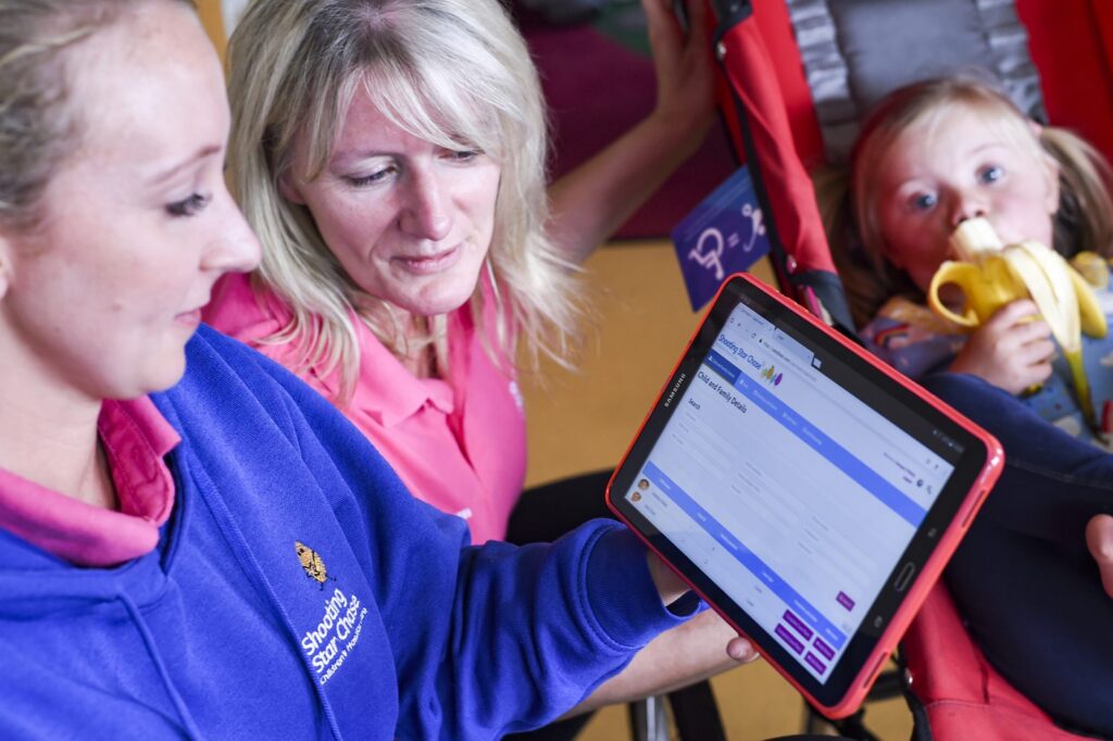 Two Shooting Star Chase hospice staff looking at a care record on a tablet, with a child in the background