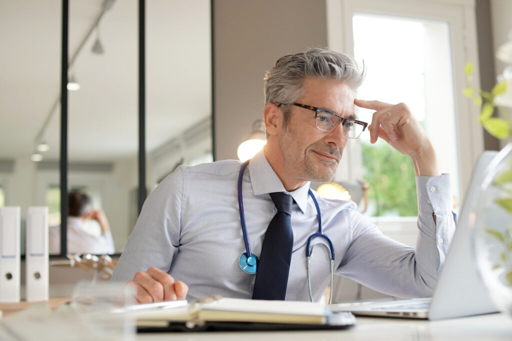 A doctor at a desk reviewing a record on a laptop